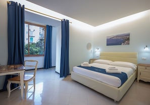 A bedroom with a bed, a desk, a chair, a window with curtains, a bedside table, a nightstand, and a wall-mounted lamp. - Sorrento Coast Relais (Meta)