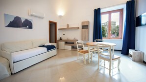 A modern room with a white sofa, a wooden dining table with chairs, a kitchen area, and a large window with curtains. - Sorrento Coast Relais (Meta)