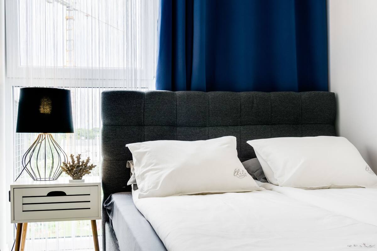 Cama bem arrumada com lençóis brancos e duas almofadas, mesa de cabeceira com abajur e um objeto decorativo pequeno, e janela com cortinas azuis.