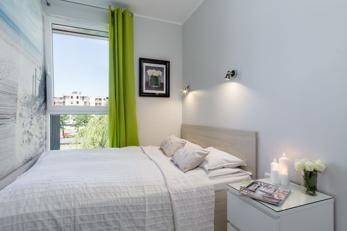 Uma cama bem arrumada com roupa de cama branca, um quadro na parede e uma mesa de cabeceira com velas e uma revista.