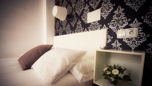 A neatly made bed with white and brown pillows, a bedside table with a vase of flowers, and a wall with a patterned wallpaper and a light fixture. - Hostal On Dormire (Valencia)