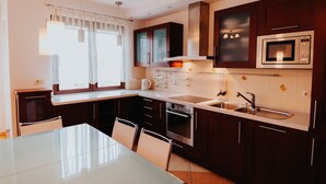 A modern kitchen with dark wooden cabinets, stainless steel appliances, a glass dining table, and a window with white curtains. - Elite Apartments Jelitkowski Dwor (Gdansk)