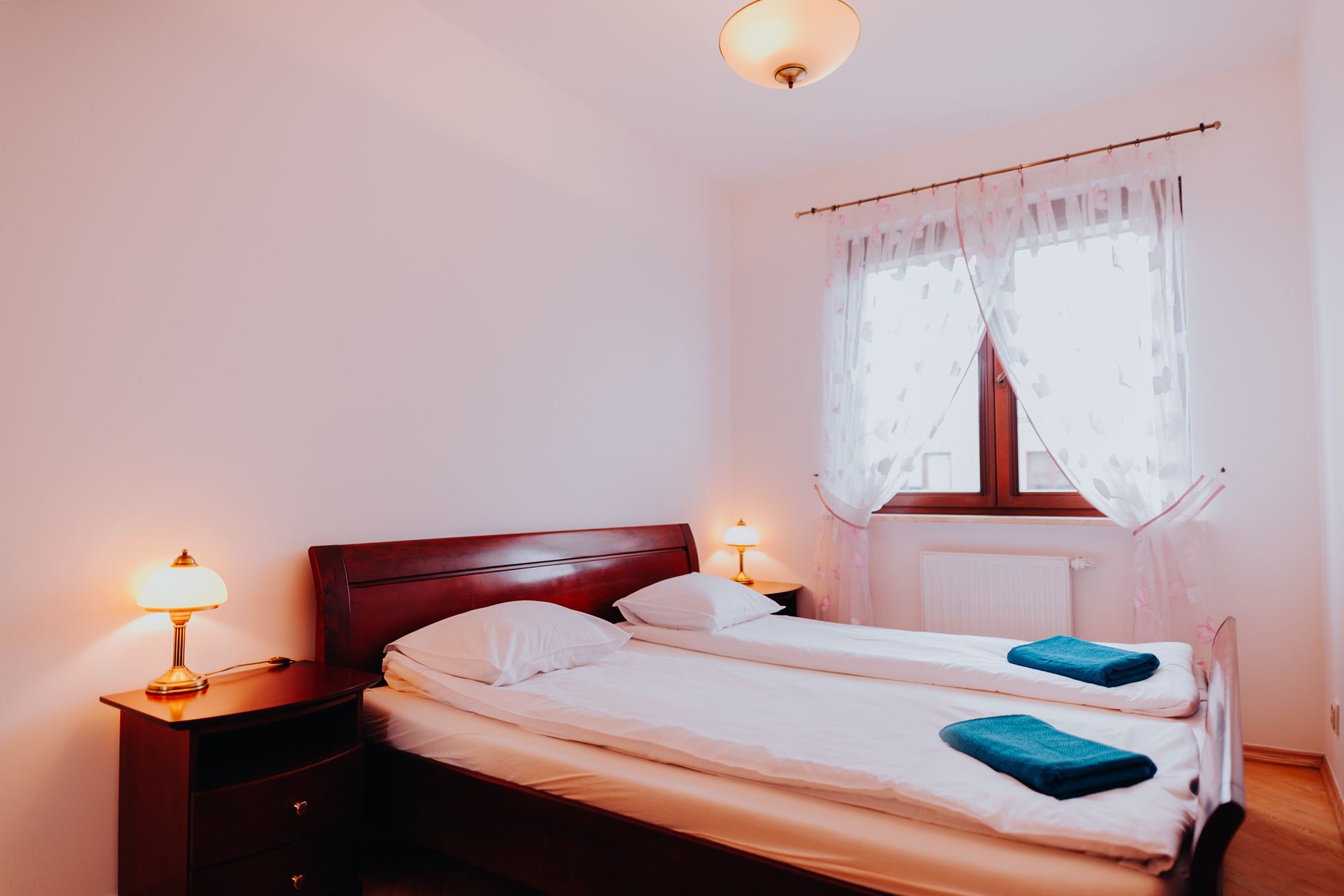 Ein Schlafzimmer mit einem hölzernen Bett, zwei Nachttischen mit Lampen, einem Fenster mit durchsichtigen Vorhängen und einer Heizung.