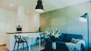 A modern kitchen with white cabinets, a dining table, and chairs. There is a blue sofa and a floor lamp in the living area. - Elite Apartments Nature & Recess (Gdansk)