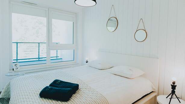 A neatly made bed with a white comforter and two pillows, a bedside table with a lamp, and a window with a view of trees. - Elite Apartments Nature & Recess (Gdansk)