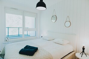A neatly made bed with a white comforter and two pillows, a bedside table with a lamp, and a window with a view of trees. - Elite Apartments Nature & Recess (Gdansk)