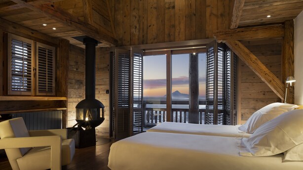 Une chambre dotée d’un grand lit, d’une cheminée et offrant une vue sur le coucher du soleil à travers des volets ouverts.