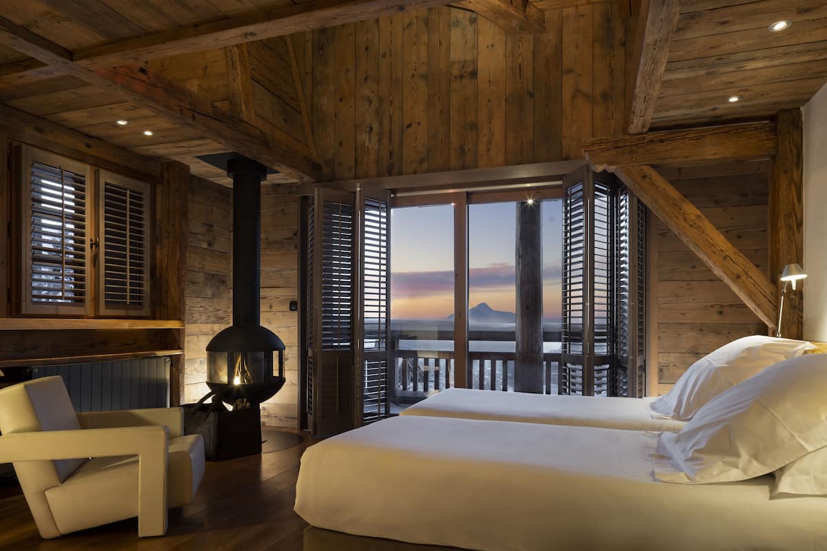 A room with a large bed, a fireplace, and a view of the sunset through open shutters.