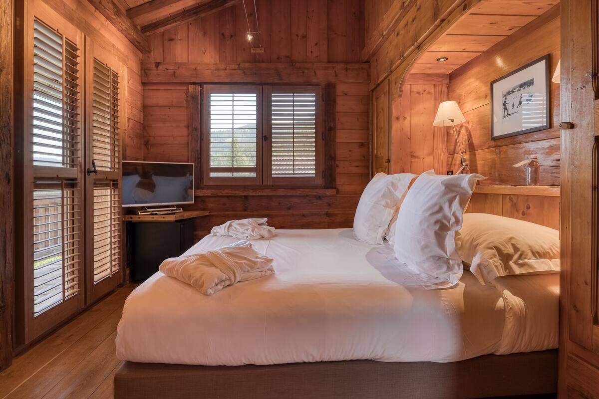 A wooden cabin room with a bed, a television, and a window with blinds.