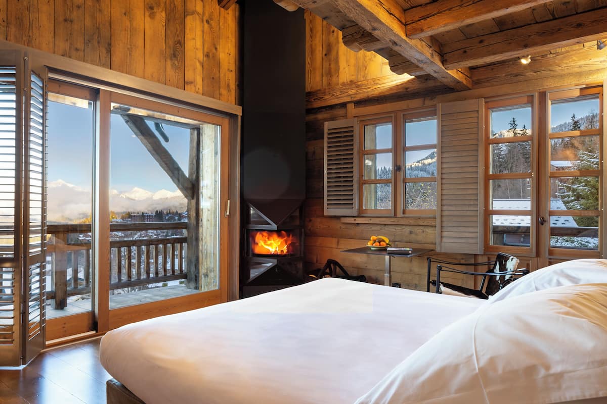 A cozy room with a bed, a fireplace, and large windows offering a view of snow-covered mountains.