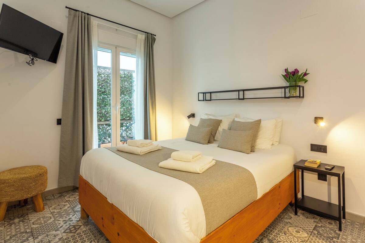 Ein ordentlich bezogenes Bett mit Kissen und Decke, ein Nachttisch mit Fernbedienung, ein kleiner Hocker, ein an der Wand montierter Fernseher und ein Fenster mit Vorhängen.