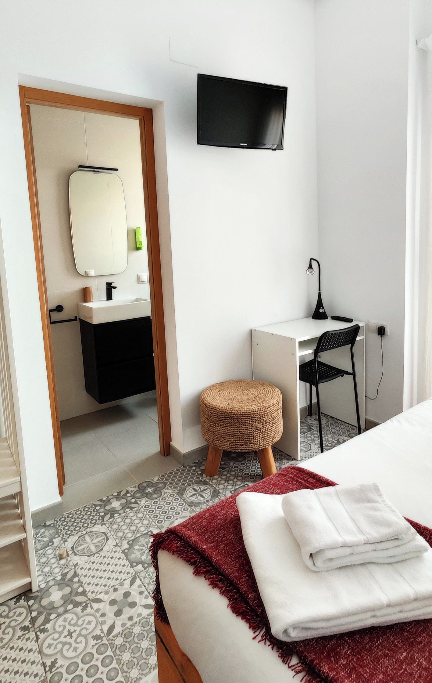 A hotel room with a bed, a desk, a chair, a TV, a mirror, and a bathroom with a sink and toilet.