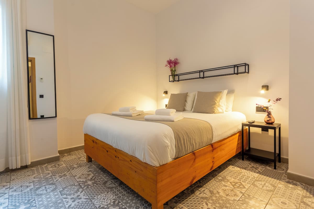 Ein ordentlich bezogenes Bett mit Holzrahmen, ein Nachttisch mit einer Vase und ein an der Wand befestigtes Regal mit Blumen.
