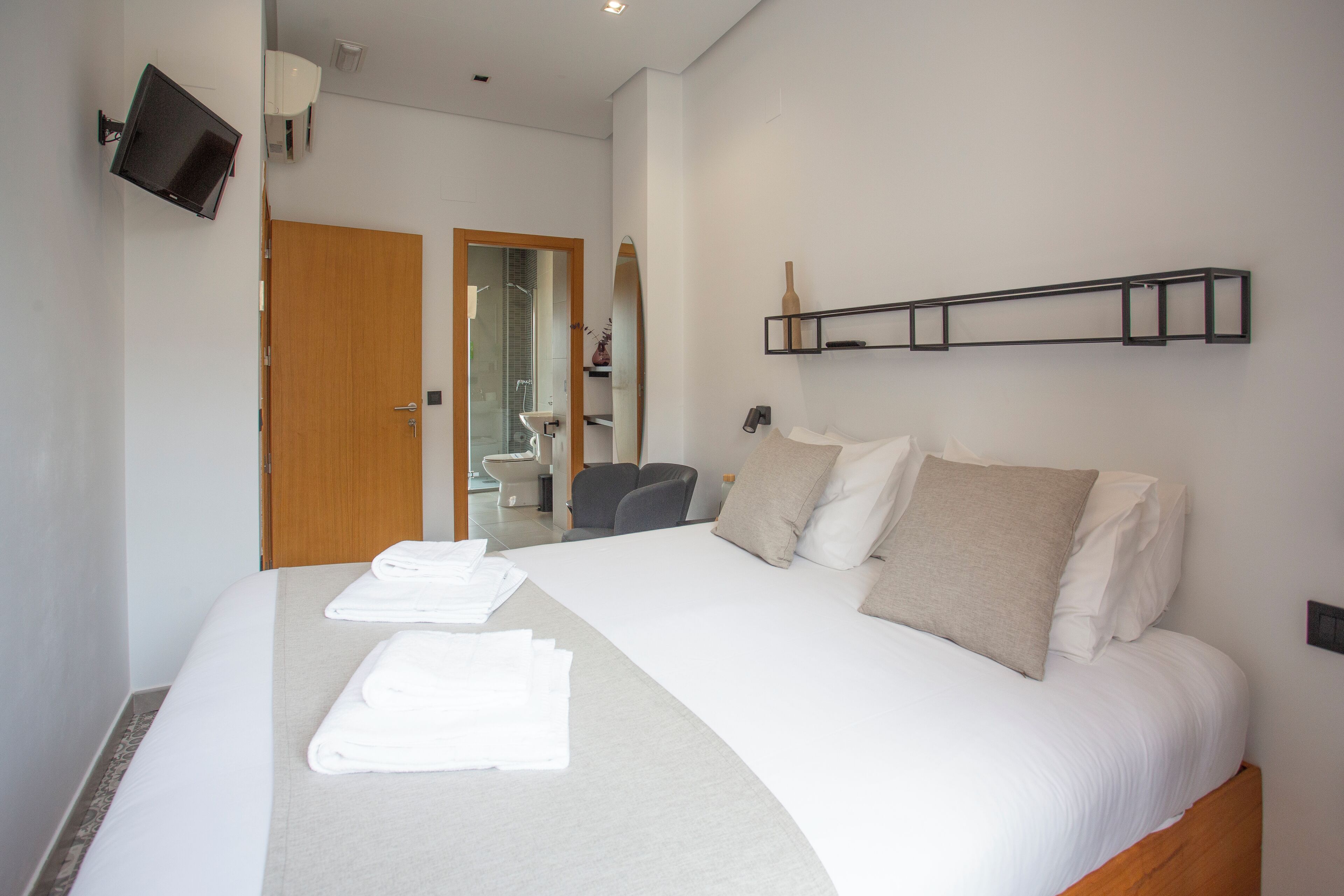 Habitación de hotel con una cama grande, un televisor de pantalla plana montado en la pared, una puerta de madera y una pequeña zona de descanso con una silla y una mesa.