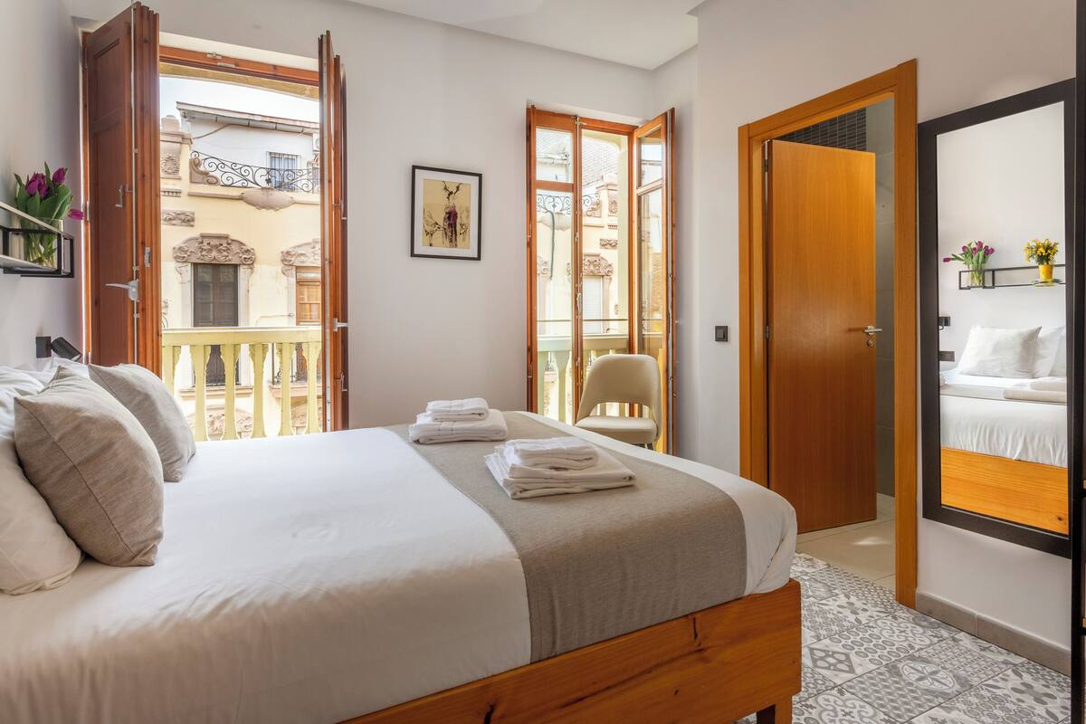 Ein Schlafzimmer mit Bett, Stuhl, einer Tür und einem Balkon mit Blick auf Gebäude.