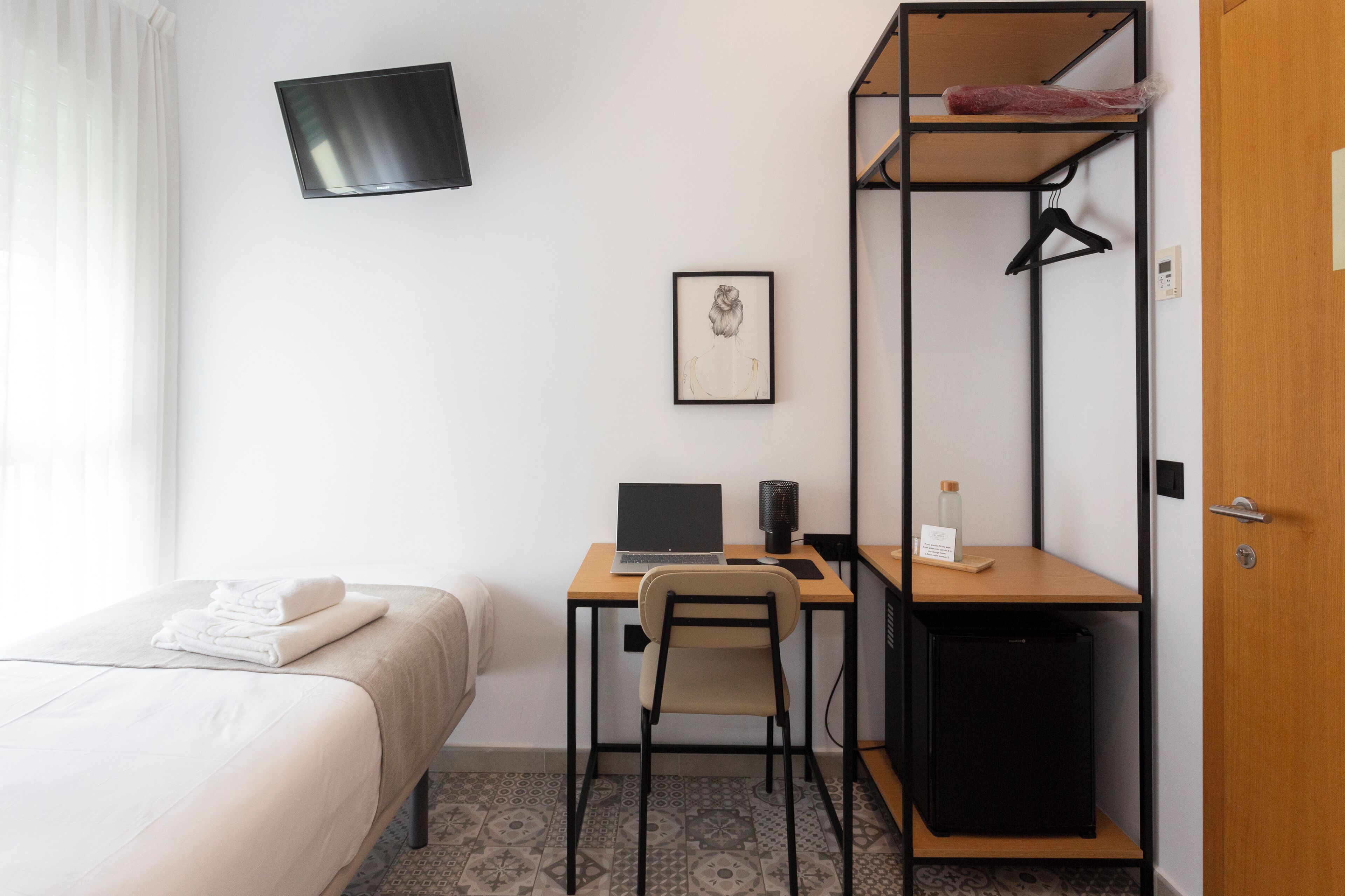 Una habitación de hotel con cama, escritorio con computadora portátil, silla, mueble pequeño, televisor montado en la pared y una puerta.