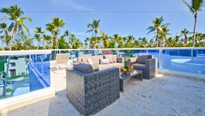 A rooftop lounge with wicker furniture, a glass balustrade, and a view of palm trees and a pool. - The Sanctuary at Los Corales (Punta Cana)
