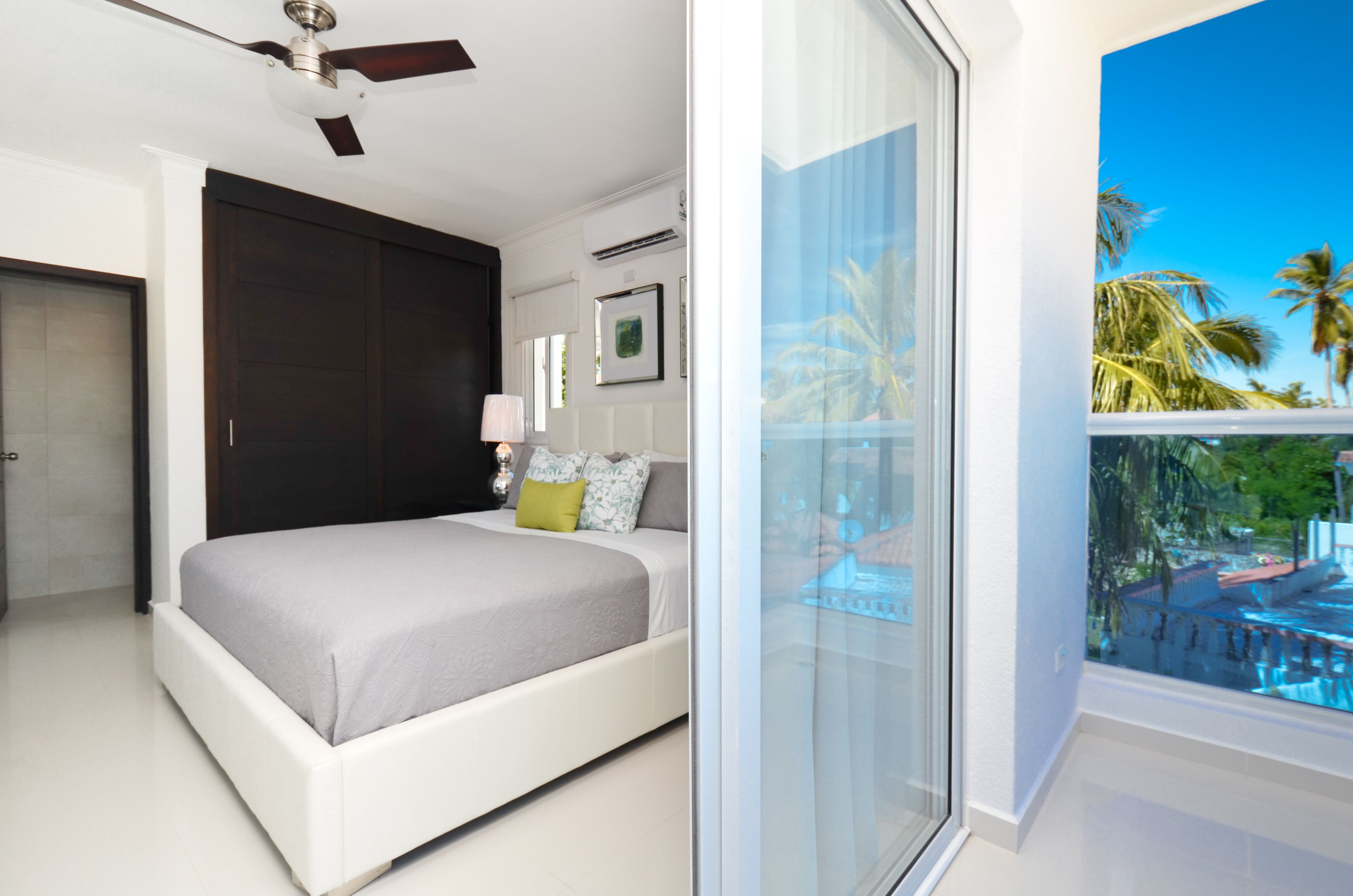 Un dormitorio con cama, ventilador de techo, ventana con vista a palmeras y una puerta.