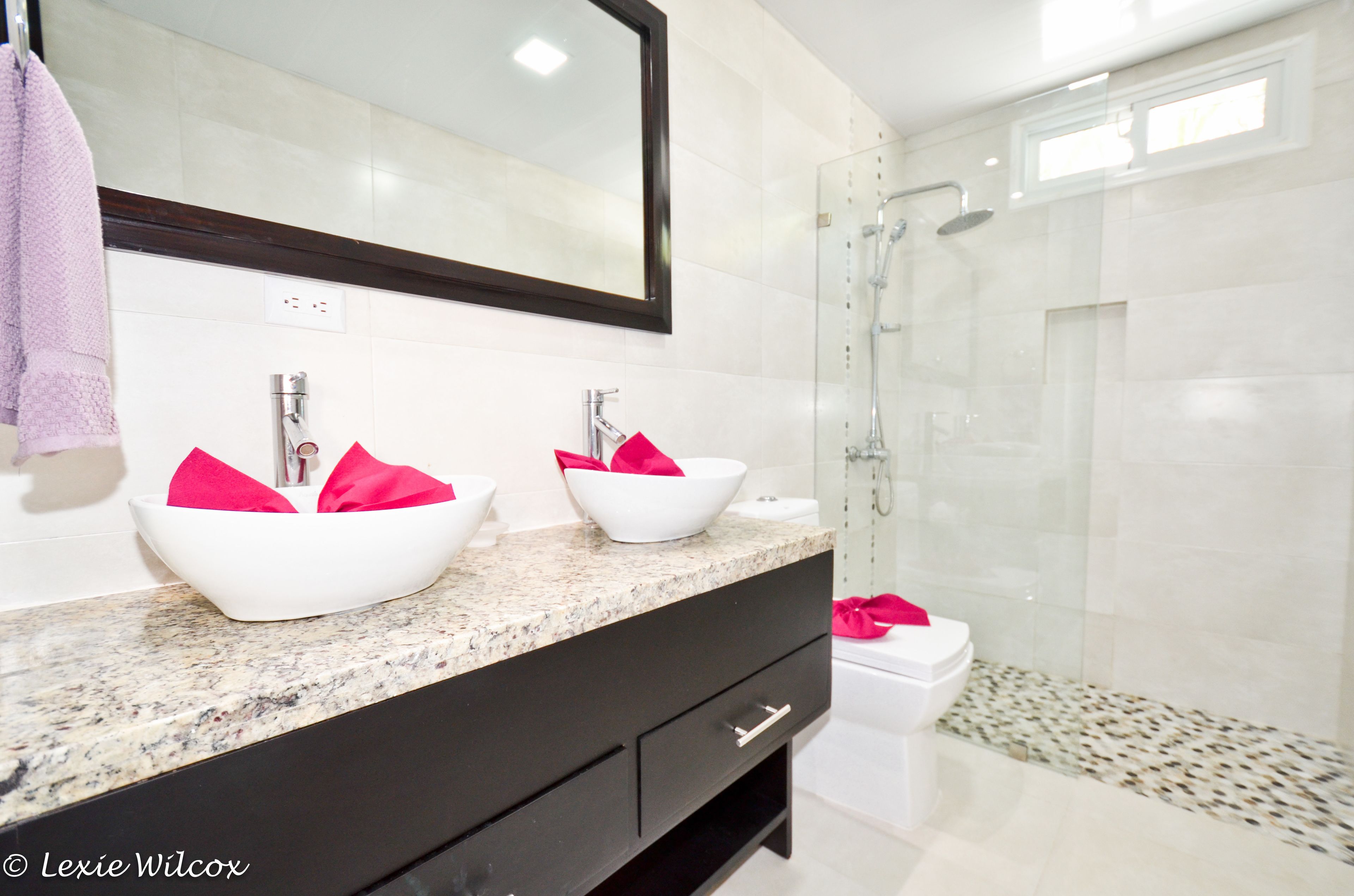 Un baño moderno con dos lavamanos blancos tipo recipiente, un espejo grande y una ducha con cerramiento de vidrio.