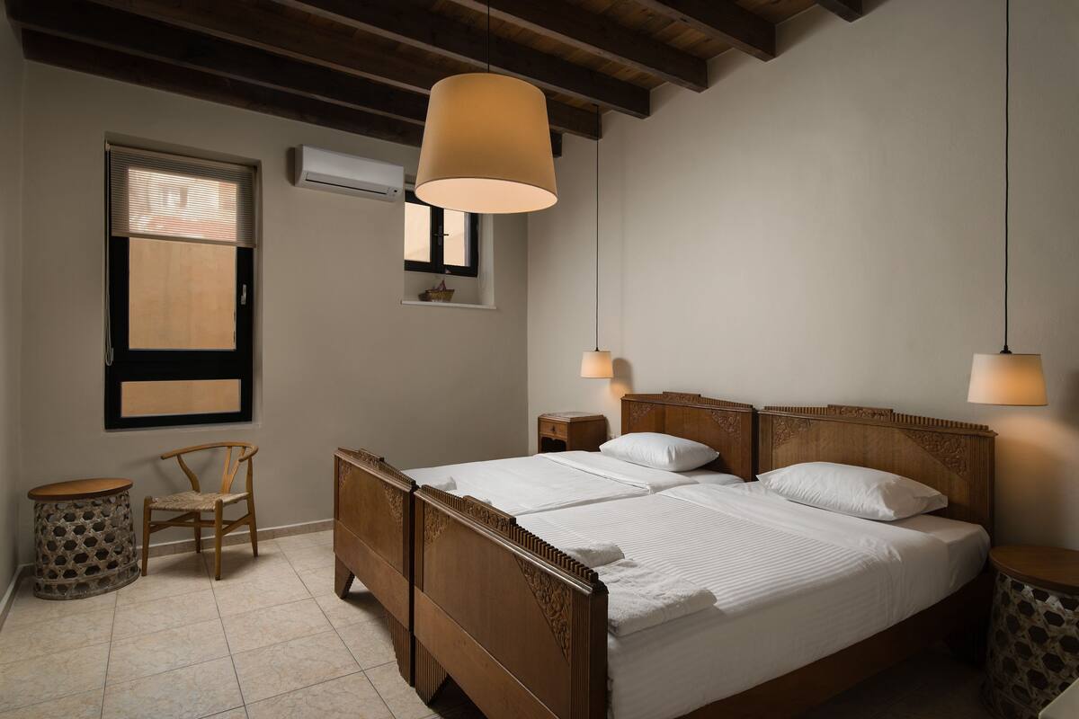 A bedroom with a wooden bed, a window with blinds, a small round table, and a hanging light fixture.