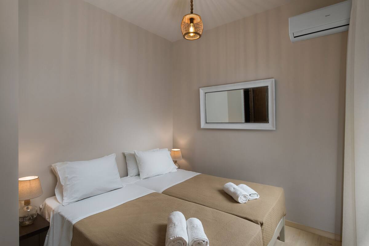 A hotel room with a bed, two pillows, a bedside lamp, a wall-mounted mirror, and a rolled towel on the bed.