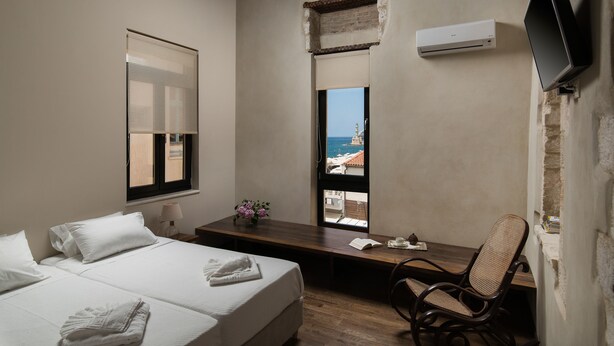 A bedroom with a bed, bedside table, chair, and a window with a view of the sea. - Nonnalena Hotel (Chania)