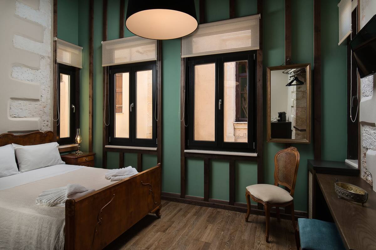 A room with a wooden bed, two windows with blinds, a chair, a desk with a mirror, and a hanging light fixture.