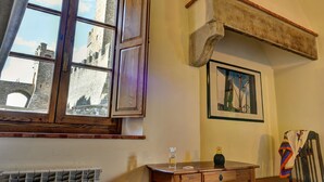 A room with a large window overlooking a historic stone tower, a radiator, a wooden table with a bottle and a vase, and a framed picture on the wall. - Albergo San Lorenzo (Poppi)