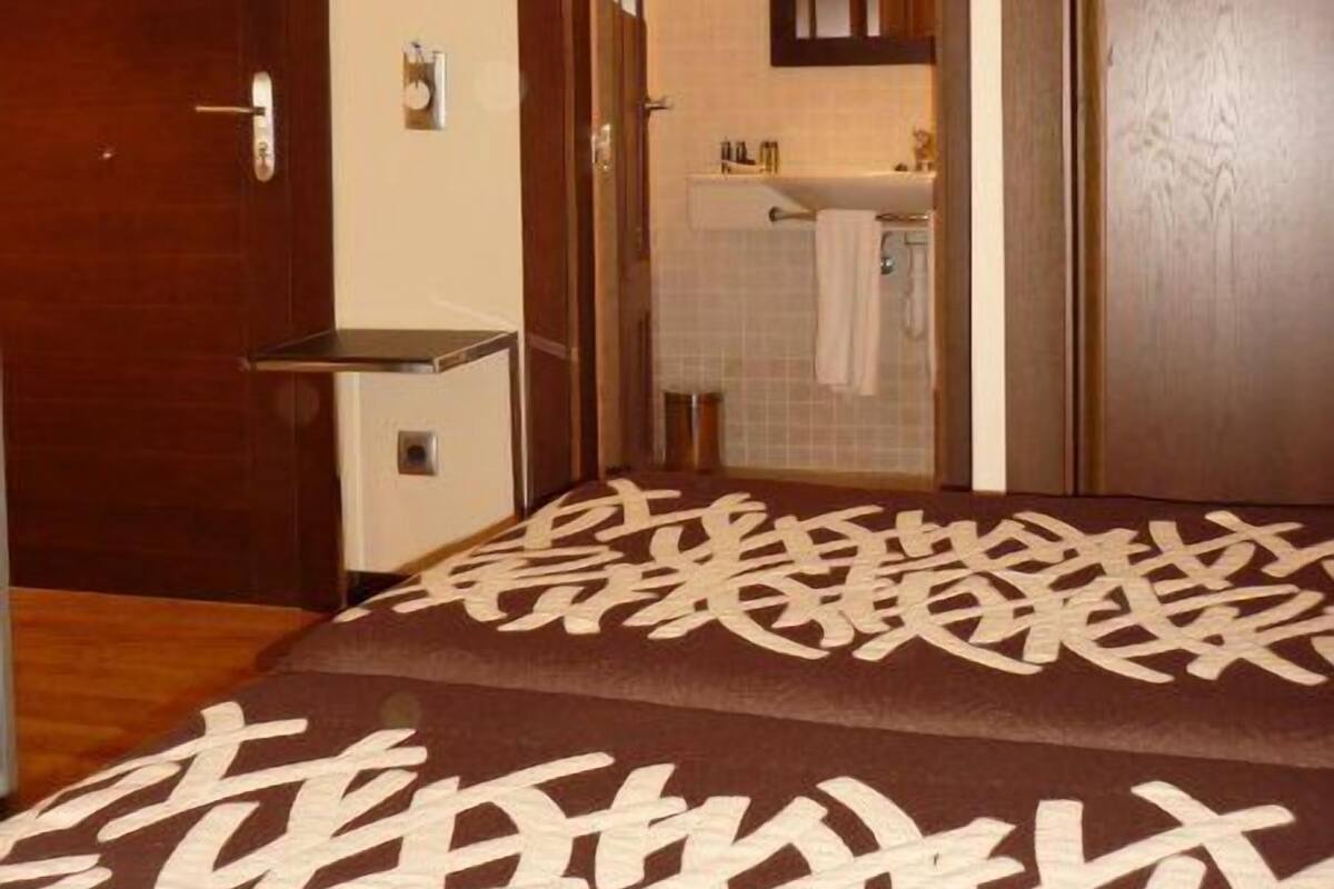 Habitación de hotel con techo de madera, cama con colcha estampada, baño con lavamanos y espejo, y puerta que da a otra habitación.