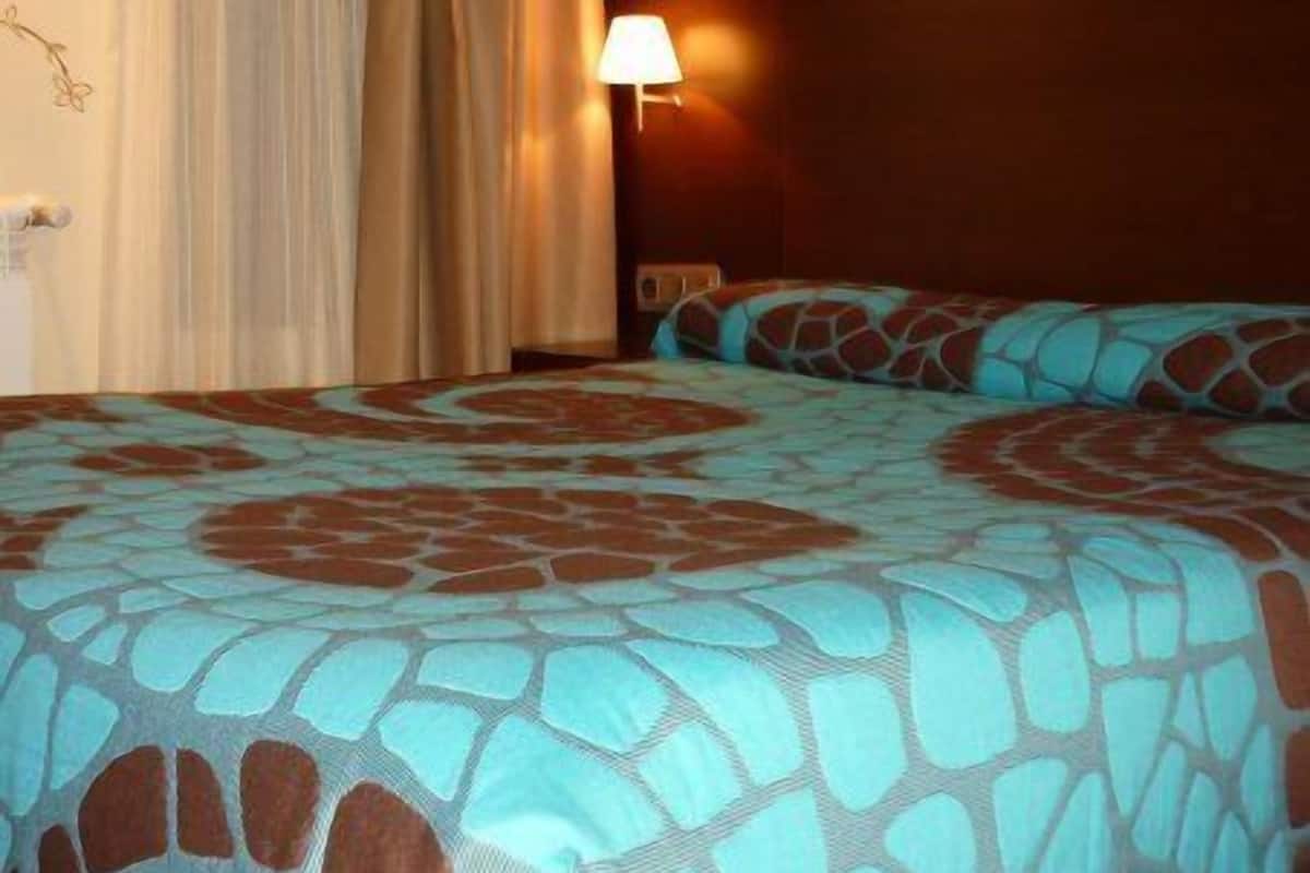 Una habitación de hotel con cama, cortinas, una lámpara de pared y una colcha estampada.