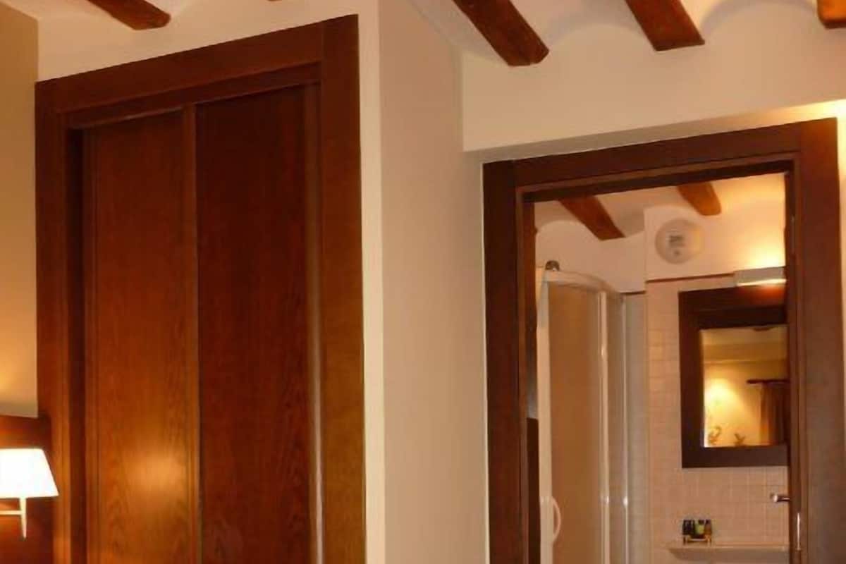 Habitación de hotel con una cama grande, vigas de madera en el techo, un baño visible a través de una puerta abierta y una mesita de noche con una lámpara.