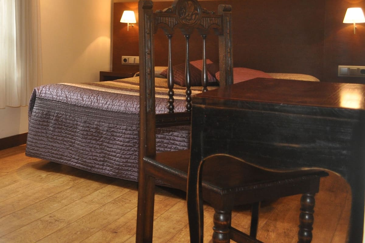 Una habitación de hotel con una cama, una silla, una mesa y dos lámparas de noche de madera.