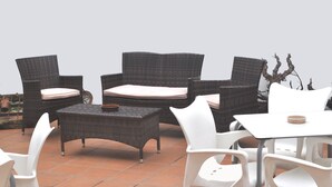 Een terras buiten met wicker meubels, witte plastic stoelen en een witte tafel.