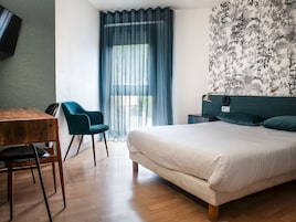 Quarto de hotel moderno com piso de madeira, cama com roupa de cama branca, uma cadeira azul, uma mesa pequena e uma TV fixada na parede.