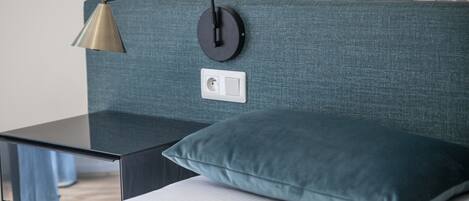 Uma cama com um travesseiro azul-esverdeado, uma mesa de cabeceira preta e uma luminária fixada na parede.