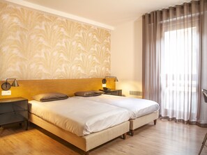 A double bed with a headboard, two bedside tables with lamps, a wall with a leaf pattern, and a window with curtains.