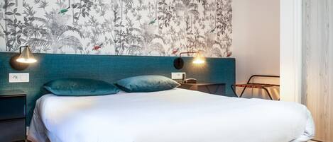 A neatly made bed with white bedding, teal pillows, and a wall with a tropical leaf pattern. There are two bedside tables with lamps and a small table with a chair.