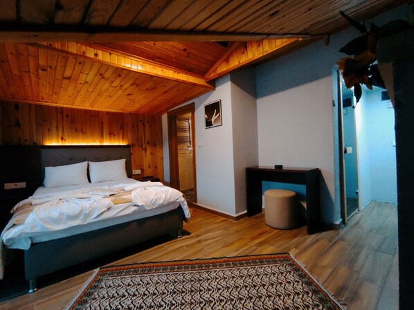 A bedroom with a wooden ceiling, a bed with white linens, a small table, and a patterned rug. - Hill River Hotel (Sile)