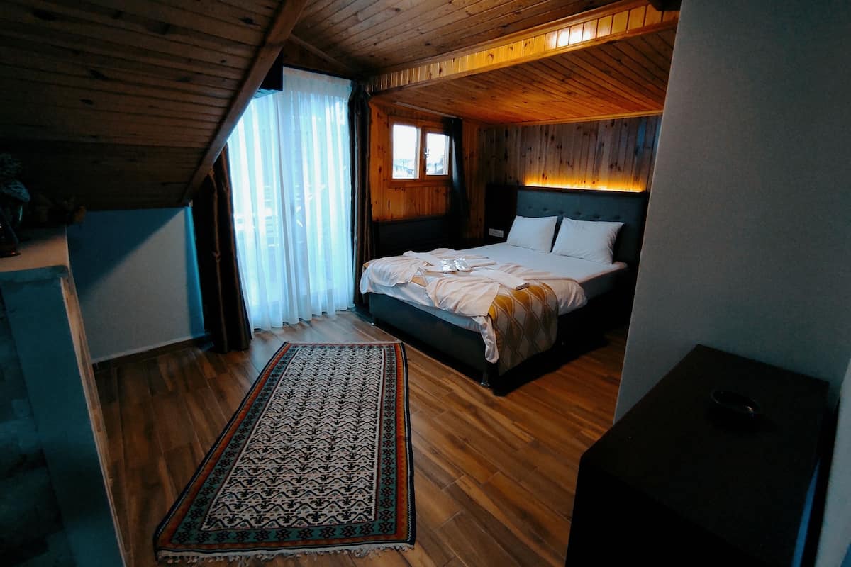 A bedroom with a wooden ceiling, a bed with white bedding, a patterned rug, and a window with curtains.
