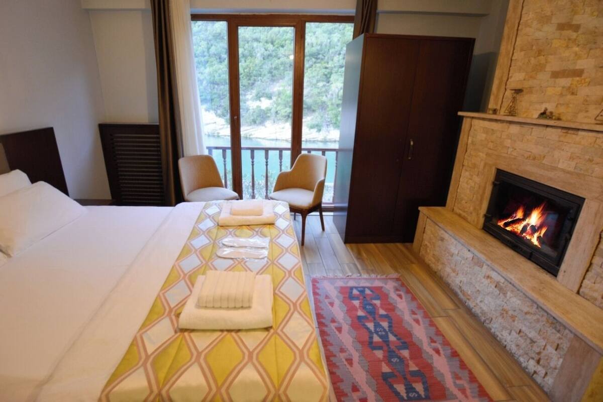A hotel room with a bed, two chairs, a fireplace, and a view of greenery through the window.