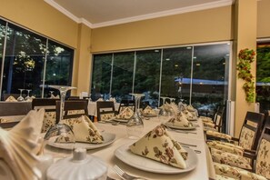 Un comedor con mesas dispuestas para una comida, ventanas amplias y sillas con tapicería floral.