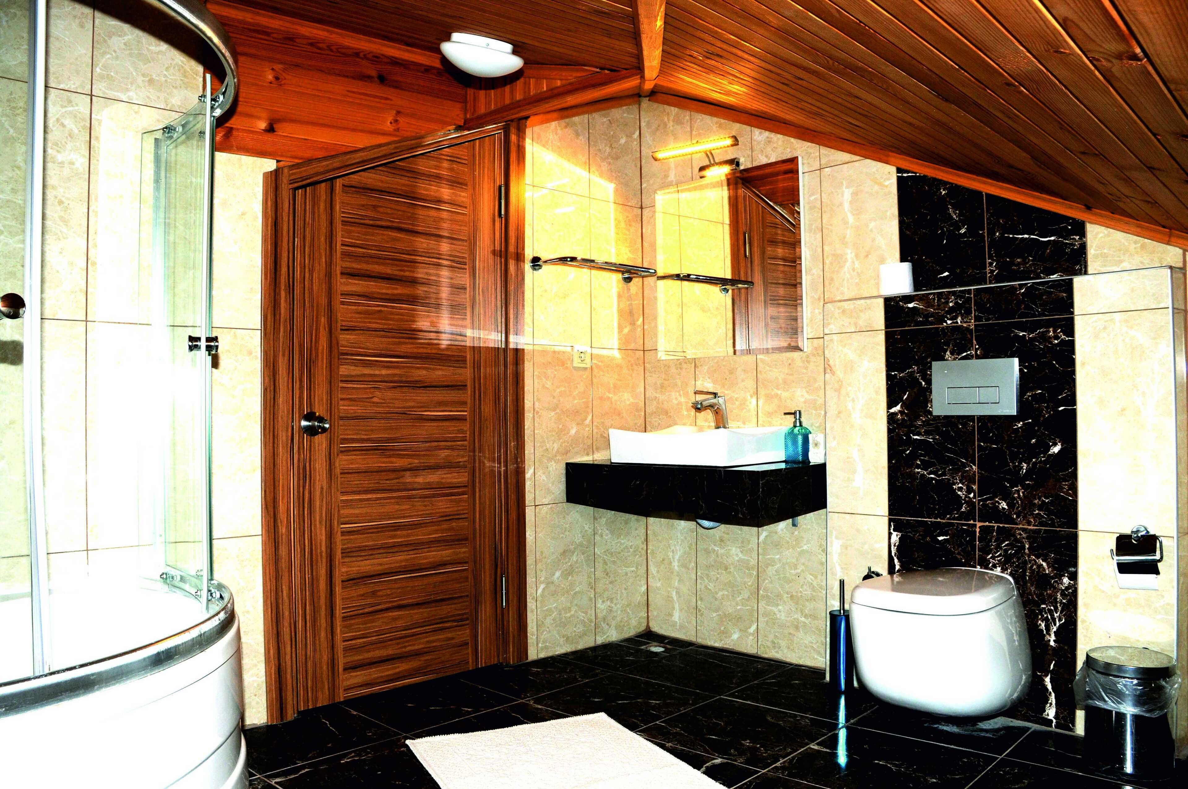 A bathroom with a wooden door, a glass shower enclosure, a sink with a marble countertop, and a toilet.