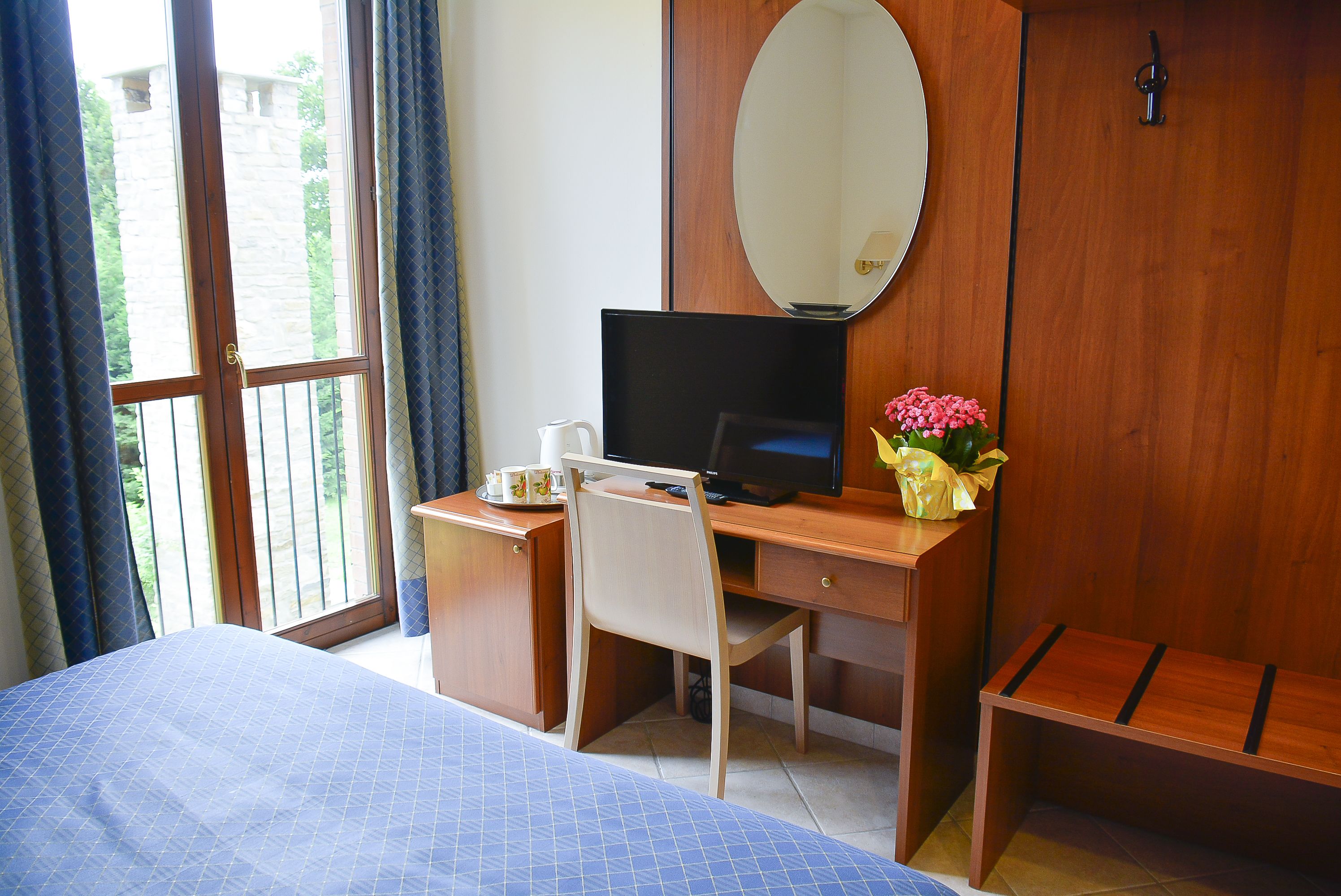 A hotel room with a bed, a desk with a computer, a chair, a wooden bench, a mirror, and a window with curtains.