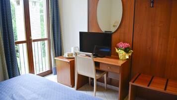 A hotel room with a bed, a desk with a computer, a chair, a wooden bench, a mirror, and a window with curtains.