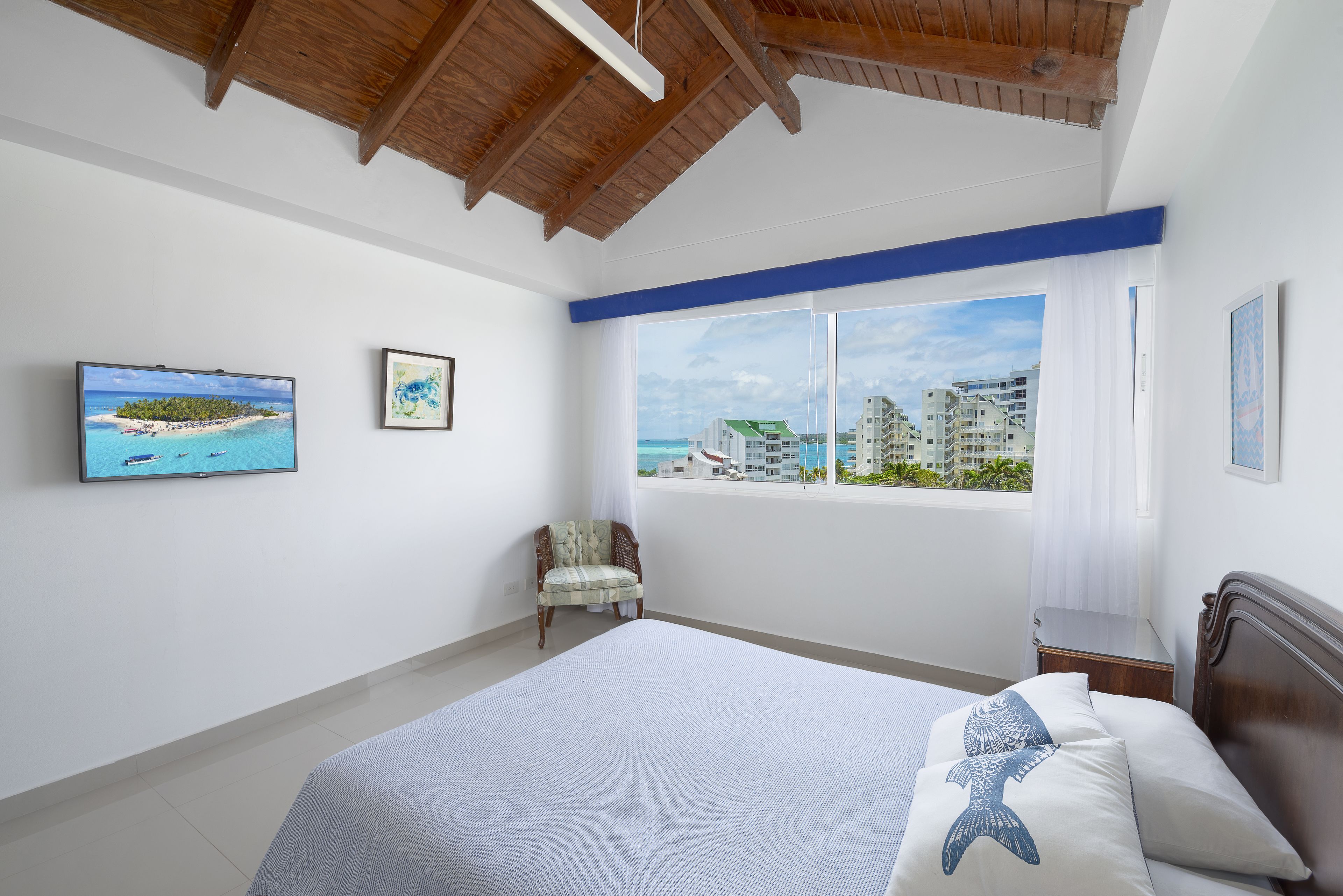 A bedroom with a large bed, a chair, and a window with a view of buildings and a beach.