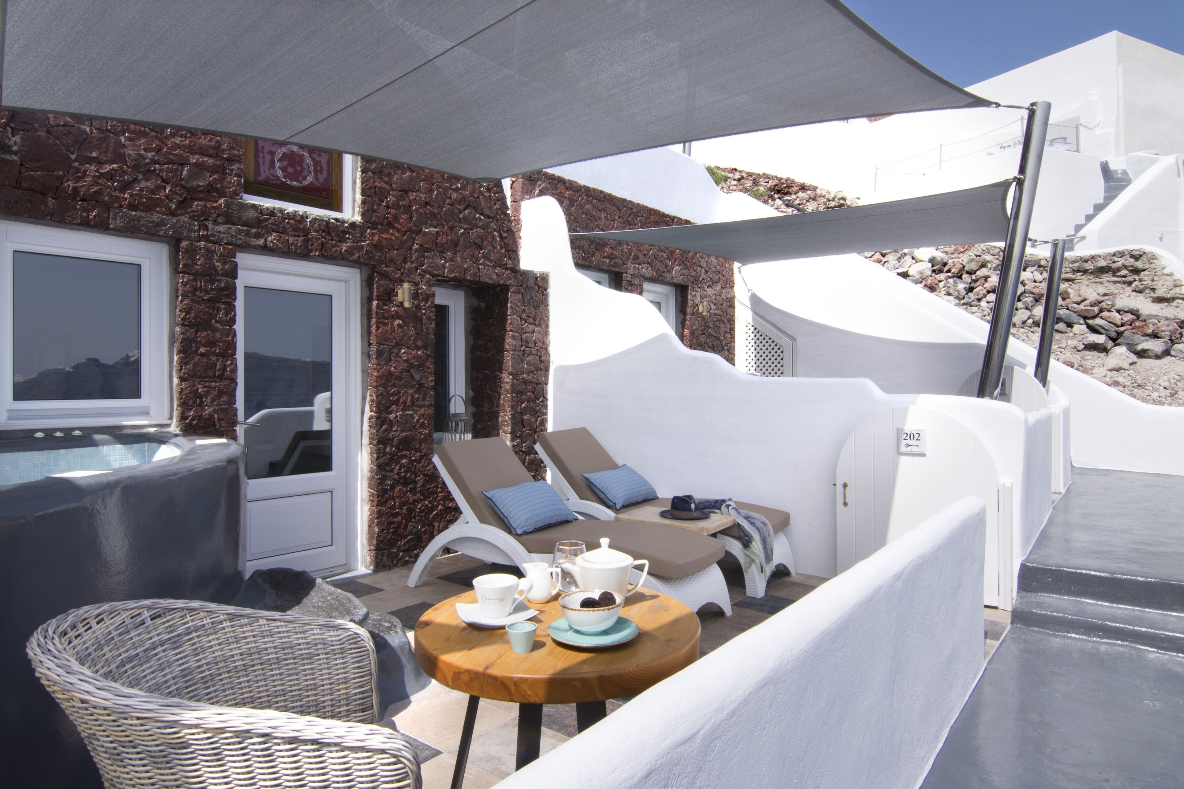 Un espacio al aire libre iluminado por el sol con sillones de descanso y una mesa lista para el té, con vistas a una piscina y un edificio de piedra.