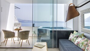 A modern room with a glass sliding door leading to a balcony, a sofa with floral cushions, two wicker chairs, a small round table with a laptop, and a large floor lamp. - Santorini Secret Premium (Santorini)