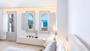 A modern bathroom with a bathtub, a bench, and three windows offering a view of the sea. - Santorini Secret Premium (Santorini)