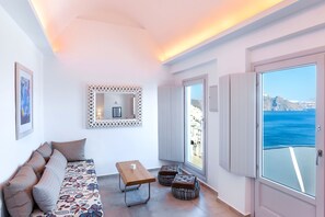 Una habitación con sofá, una mesa, otomanas, un espejo y un amplio ventanal con vista al mar.