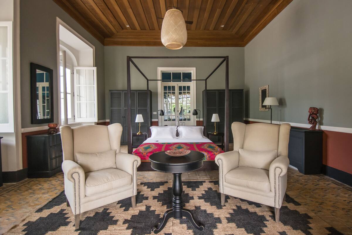 A bedroom with a four-poster bed, two armchairs, a round table, and a chandelier.